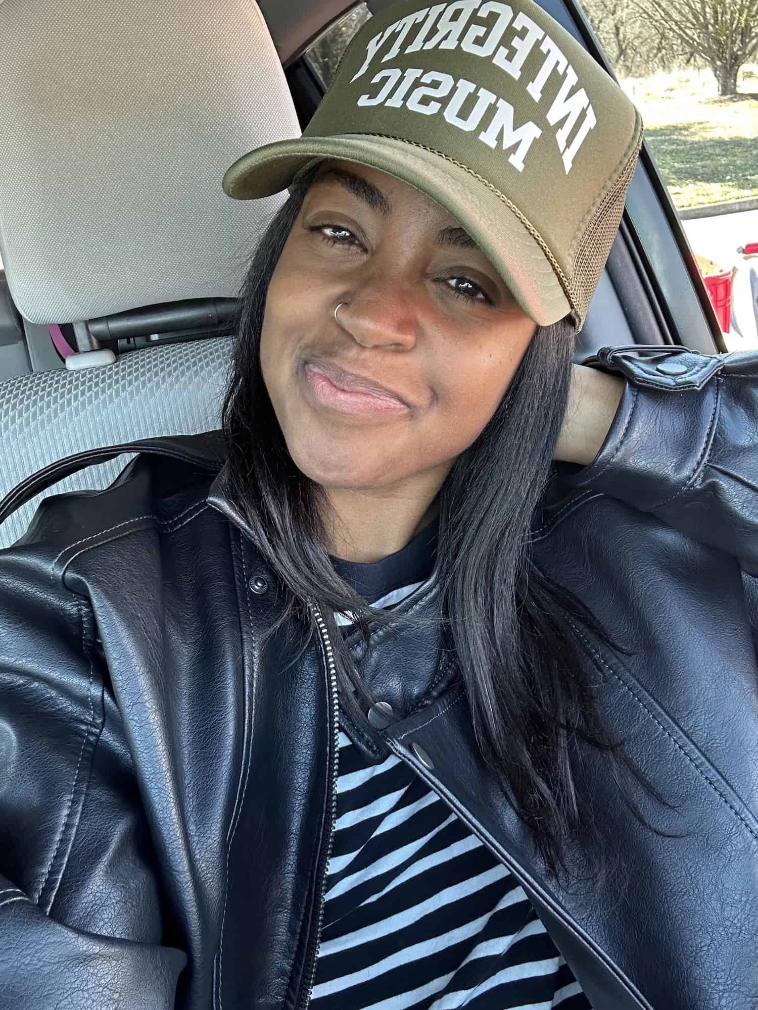 Abby Flynn wearing a "Integrity MUSIC" cap and black leather jacket, sitting in a car.